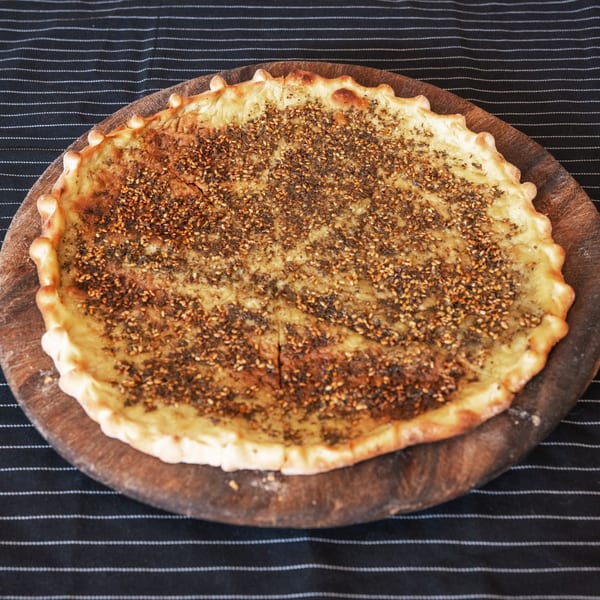Moustache Pitza's za'atar manakish, a round flatbread topped with herbs and sesame seeds on a wooden board.