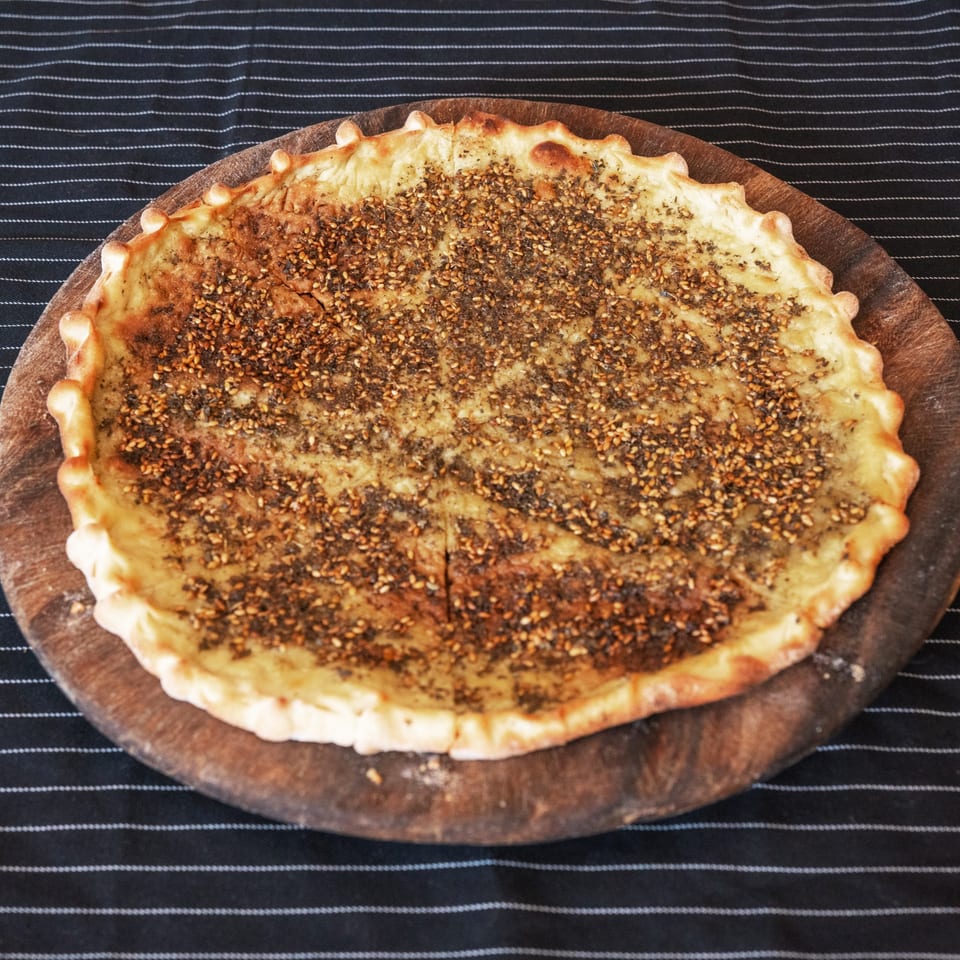 Moustache Pitza's za'atar manakish, a round flatbread topped with herbs and sesame seeds on a wooden board.