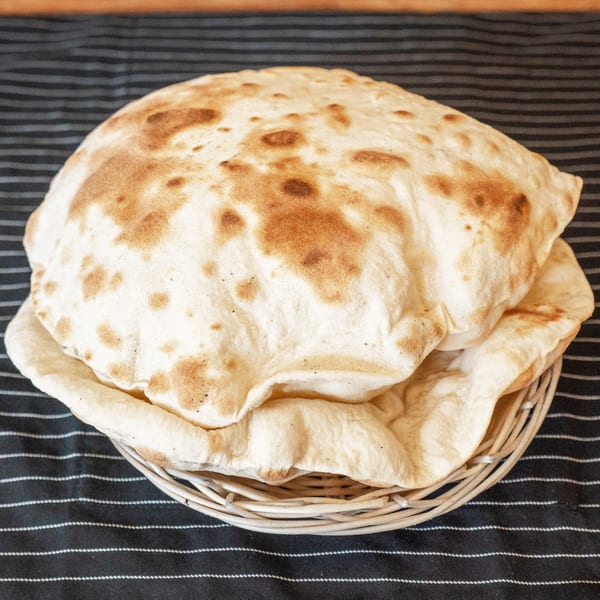 Freshly baked, golden-brown pita bread in a woven basket on a striped tablecloth.